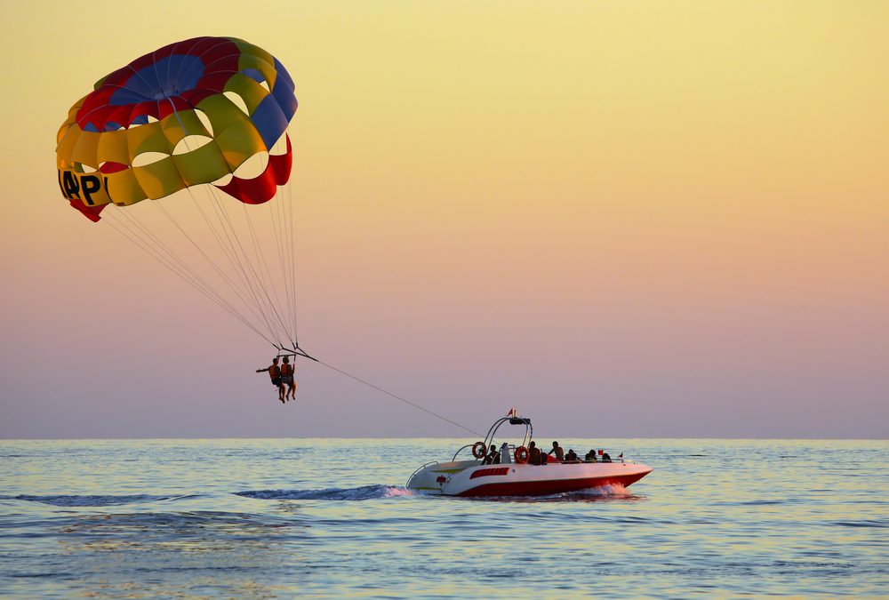 Two parasailers under a colorful canopy at sunset, towed by a boat over the Gulf